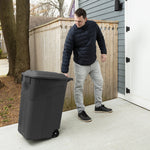 Lifestyle photo of a person tilting a black Hyper Tough wheeled trash can outdoors next to a house and fence.