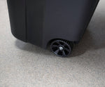 Close-up of a black plastic wheel and lower corner of a wheeled outdoor trash can on a textured floor.