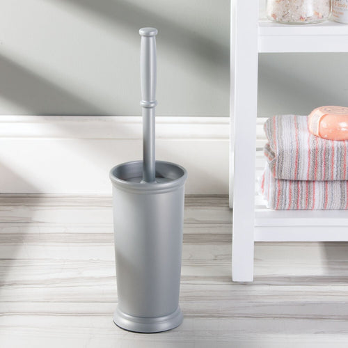 Silver cylindrical toilet brush holder with matching handle placed on a bathroom floor next to shelving and towels.