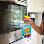 Person holding a bottle of Invisible Glass cleaner in front of a microwave, ready to spray with a yellow trigger nozzle