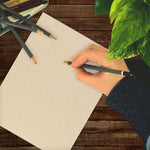 Hand writing on a natural parchment sheet on a wooden surface with pencils and a leafy plant visible
