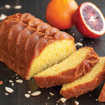 Sliced golden loaf cake with decorative patterned top on a dark board, with halved blood orange and scattered almond slices.