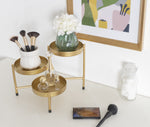 Staged tabletop scene with a gold tri-level plant stand holding decorative vases and perfume bottles beside framed art