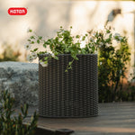 Keter logo above a graphite woven-style planter holding white flowers on a wooden outdoor surface