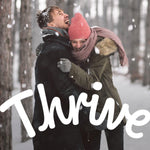 Couple embracing and laughing in falling snow with large 'Thrive' branding overlay, lifestyle winter scene