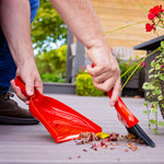 Kneeling person sweeping small garden debris with a red whisk broom into a matching dustpan