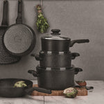 Styled kitchen scene with stacked speckled saucepans and lids, hanging frypans, herbs, and a cutting board.