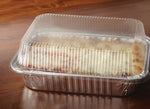 Cooked lasagna inside a foil aluminum pan topped with a clear ribbed snap-on plastic lid on a wood surface.