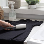 Hand using a lint roller with dark gray handle on a black T-shirt on a countertop with household items in background