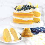 Sliced layered lemon cake with white frosting, blueberries and lemon wedges displayed on a white pedestal cake stand.