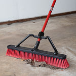 Libman red-bristled push broom scraping leaves and dirt from a concrete floor, angled close-up