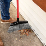 Libman angle broom being used to sweep leaves and debris along a house siding edge, showing red handle and gray bristles
