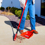 Person using a red Libman dustpan and broom outdoors, broom sweeping debris into the scoop near pavement.