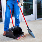 Person using a red-handled broom and black upright scoop dustpan to sweep outdoor debris on concrete.