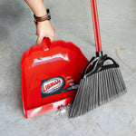 Hand holding a red Libman dustpan on a concrete floor next to a broom, label visible inside the scoop.
