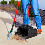 Person standing while using a red-handled broom with a black upright scoop dustpan holding debris on pavement.