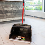 Black Libman upright scoop dustpan with red handle on a tiled floor near a counter, label visible inside pan.