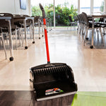 Libman black upright scoop dustpan with red handle placed on a tiled floor in a room with tables and chairs.
