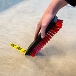 Close-up of a hand holding a Libman black resin scrub brush with red bristles scrubbing a concrete surface