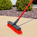 Red-handled Libman push broom sweeping a textured outdoor concrete surface with landscaping in background