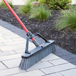 Libman push broom in use outdoors sweeping pavers, showing the red handle, black support bracket, and gray bristles