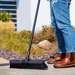 Lifestyle photo of Libman push broom on patio pavers with handle and a person's lower legs and boots visible