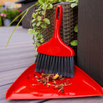 Red Libman whisk broom with black bristles resting in a red dustpan beside a wicker planter