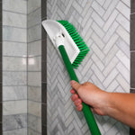 Hand holding a Libman green-handled scrub brush cleaning grout and tile on a herringbone-pattern shower wall