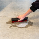 Hand-held Libman scrub brush with black ribbed handle and red bristles being used on a wet concrete floor