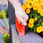 Hand using a red Libman scrub brush to clean the edge of an outdoor planter near yellow flowers