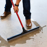 Person using a red-handled squeegee to push water across a concrete floor, showing blade contact and footwear