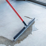 Red-handled floor squeegee pushing a puddle of water across a smooth concrete surface near a window