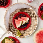 Lifestyle overhead scene with small red plate holding watermelon and blueberries nested in woven tray on white table