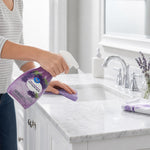 Person spraying a Great Value lavender multi-purpose cleaner onto a white bathroom countertop, with faucet and lavender sprig nearby.