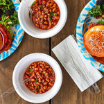 Top-down lifestyle scene with bowls of chili, paper plates, a burger and clear plastic forks and spoons on a wooden table