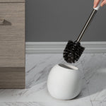 Hand lifting black-bristled brush from a white rounded dome holder beside baseboard and cabinet