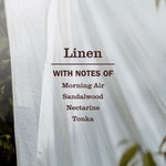 Close-up of a white linen sheet hanging with text listing scent notes: Morning Air, Sandalwood, Nectarine, Tonka.