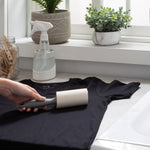 Lifestyle scene of a hand using a lint roller on a black t-shirt on a countertop near plants and spray bottle