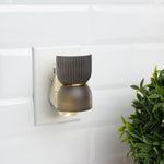 Ribbed dark gray fragrance plug lit and plugged into a wall outlet beside a decorative green plant
