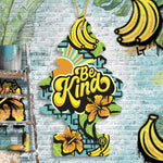 Be Kind tree-shaped air freshener hanging against a graffiti-style brick wall with bananas and floral artwork.