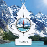 Close-up of a Little Trees True North tree air freshener with compass motif over a snowy mountain and lake background