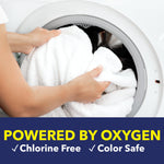 Hands loading white towels into the drum of a front-loading washing machine with banner text stating 'Powered by Oxygen' and product claims.