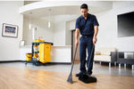 Worker using a tall black upright dust pan and broom on a wood-look floor in a lobby setting with janitorial cart in background.