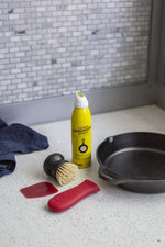 Staged kitchen scene showing Lodge seasoning spray, scrub brush, red silicone tools and a cast iron skillet on a light countertop near tiled backsplash.