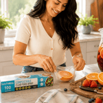 Person stretching clear cling wrap over a halved orange with Lunchskins compostable clear food wrap box on counter