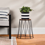 Styled room scene showing the acacia wood top plant stand with black wire base holding a striped planter beside a sofa