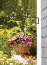 Black metal plant hanger bracket mounted on a white siding wall holding a hanging basket with pink flowers in a garden setting