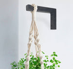 Black L-shaped wooden wall bracket mounted on a white wall holding a macramé plant hanger with green leaves.