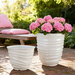 Two cream ribbed planters outdoors, one filled with pink hydrangeas beside a wooden bench