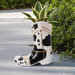 Ceramic cowboy boot planter with black spots holding a variegated plant, outdoor staged scene